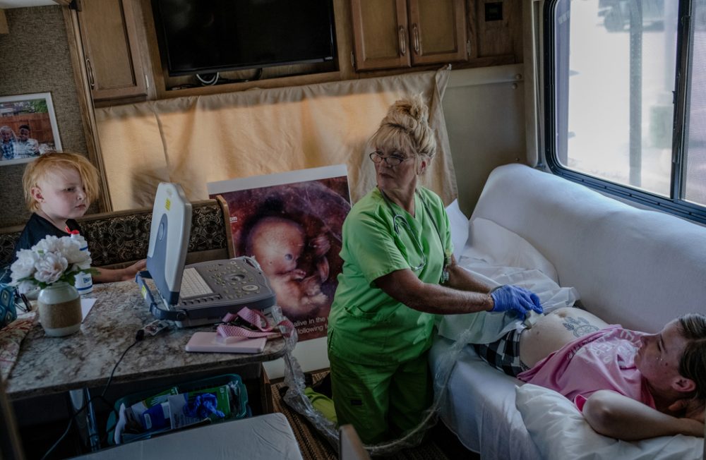 Angela Michael (67) (center), an anti-abortion activist and former nurse, performs ultrasounds in a recreational vehicle transformed into a small clinic outside the Hope Clinic for Women in Granite City, Illinois. Michael and her husband park their RV outside of the abortion clinic nearly every day to dissuade patients from seeking abortions. They offer diapers and other supplies instead. Years earlier, she convinced Ashley Miller (29) (right) not to have an abortion. Ashley is now five weeks into her 4th pregnancy, likely with twins. She had a miscarriage a few months before after being beaten by her partner, a local police officer. In the first nine months after the Supreme Court's ruling, Illinois saw an increase of 12,400 abortions from the previous year as people traveled there from states with bans, according to the Society of Family Planning. The Michaels are concerned about this surge. Granite City, Illinois, USA, 19.07.2022  (The scene was not staged; permission was granted by Angela and Ashley, her daughter's legal guardian.)