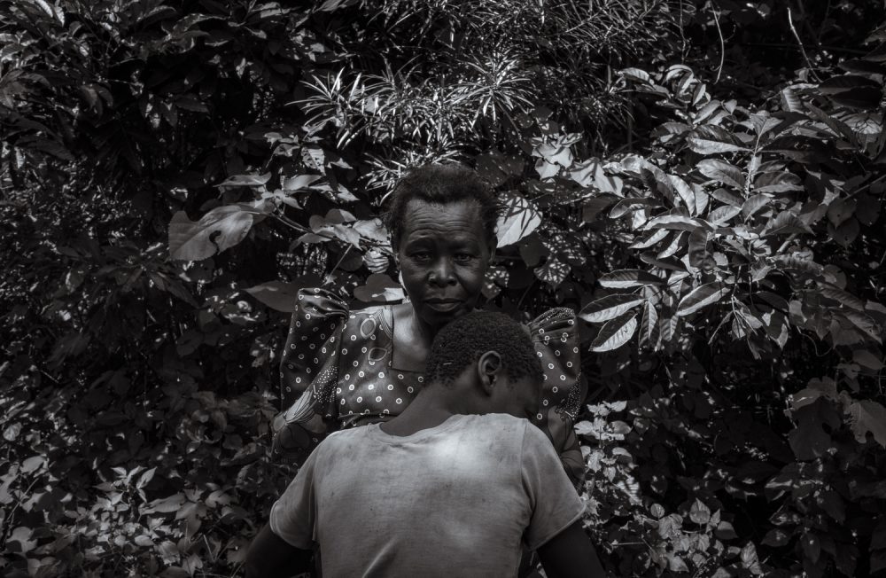 19 July 2023. Nabwigulu, Kamuli District, Uganda. Perez Mbwase cuddles into his mother Lovinsa Namwebya's bosom after being admonished for misbehaving. 
Due to his Autism, hunger is a trigger for Perez' mood to swing towards aggression. After walking for over two hours so Lovinsa could inspect a potential plot of land to plant vegetables Perez didn't eat, causing him to act out. Namwe tried for years to have her son’s condition diagnosed. Unable to deal with his aggression and hyperactivity, his parents tied him to a tree for 11 years. He was fed and toileted in the same place until late evening when he would join the family inside. Following the death of his father from suicide, Lovinsa struggled to care for Perez alone and he was moved to the Amaanyi Centre. While there he was formally diagnosed with severe, category four, Autism. During his time at the Amaanyi Centre Perez would learn basic skills such as dressing himself and eating with cutlery. Almost as importantly, the staff would educate Lovinsa around Autism. She would be taught what Autism is, how it manifests in Perez and what his triggers are. This education would lead to a tighter bond between mother and son and provide Lovinsa with the tools she needed to properly care for her son.